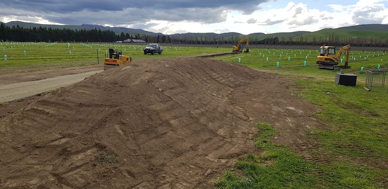 Heavy machinery working in a field. Brown dirt in foreground, with trees and hills in the background. Overcast sky.