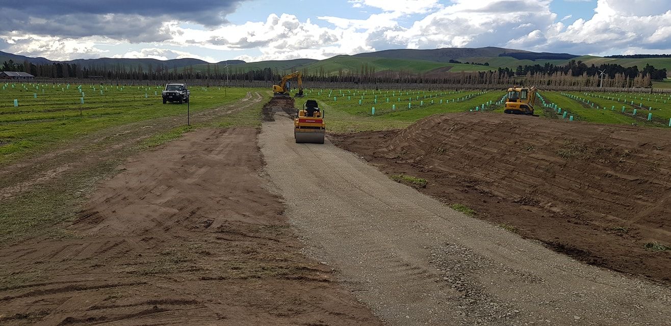 Road construction in a field, with heavy machinery including a roller and an excavator. Cloudy sky, rolling hills.