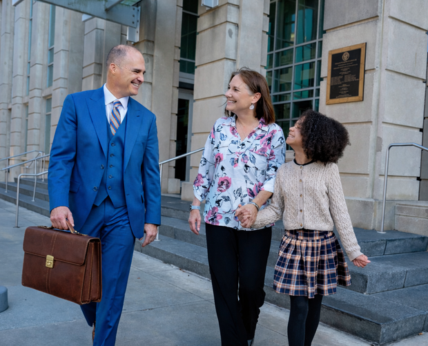 Man in blue suit, woman, and girl walking past a building; smiling. Man carries a briefcase.