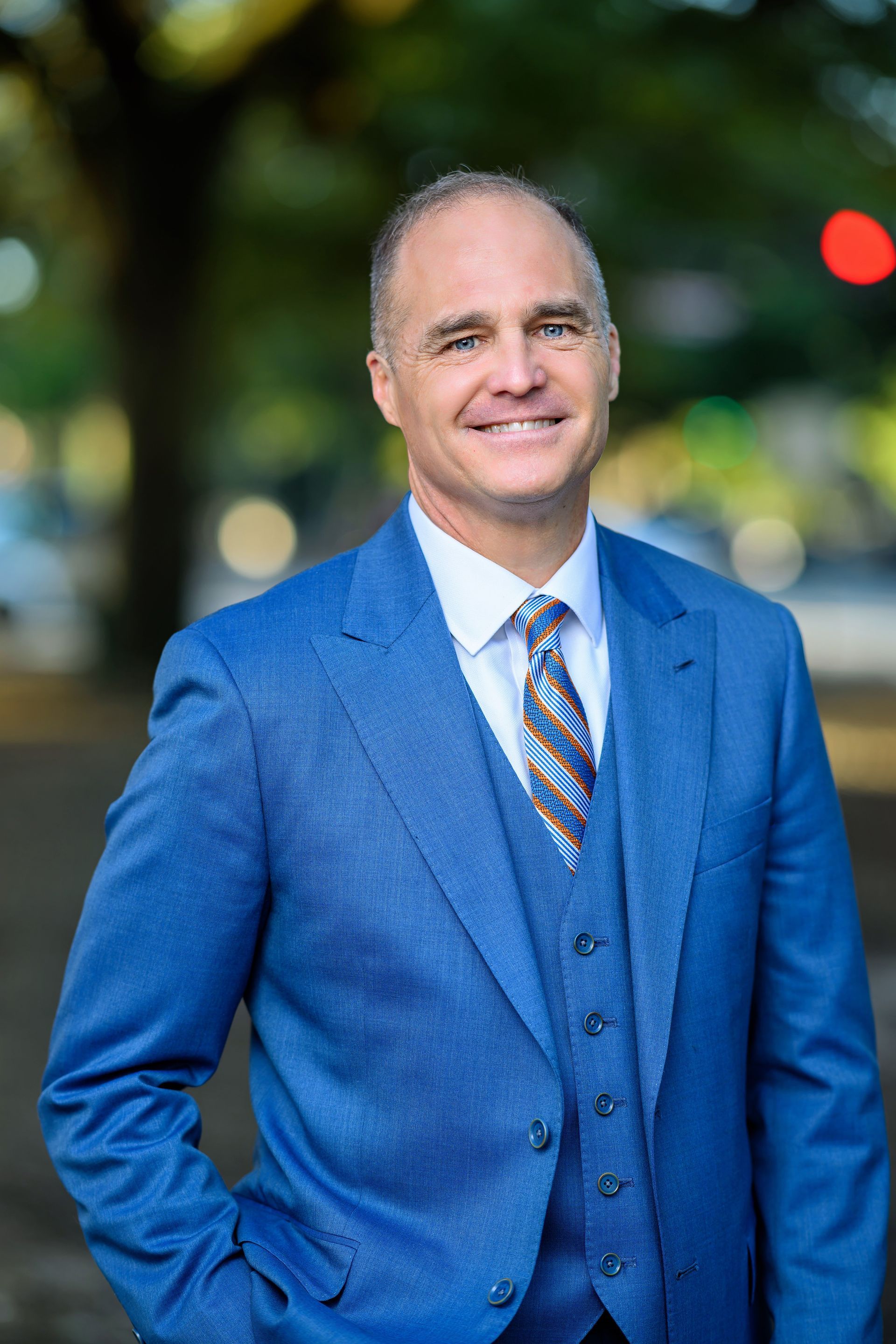 Man in blue suit, smiling outdoors with a city background.