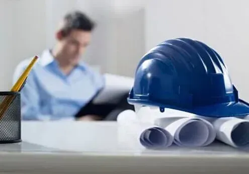 Blue hard hat and rolled blueprints on a desk, with a blurred person working in the background.