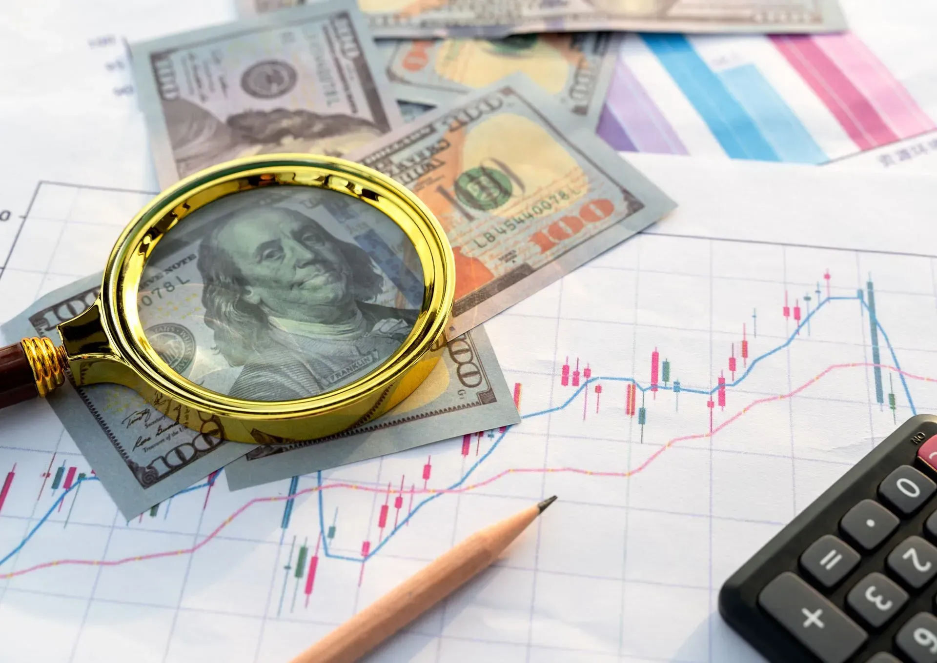 Magnifying glass over dollar bills and stock chart with calculator and pencil, showing financial analysis