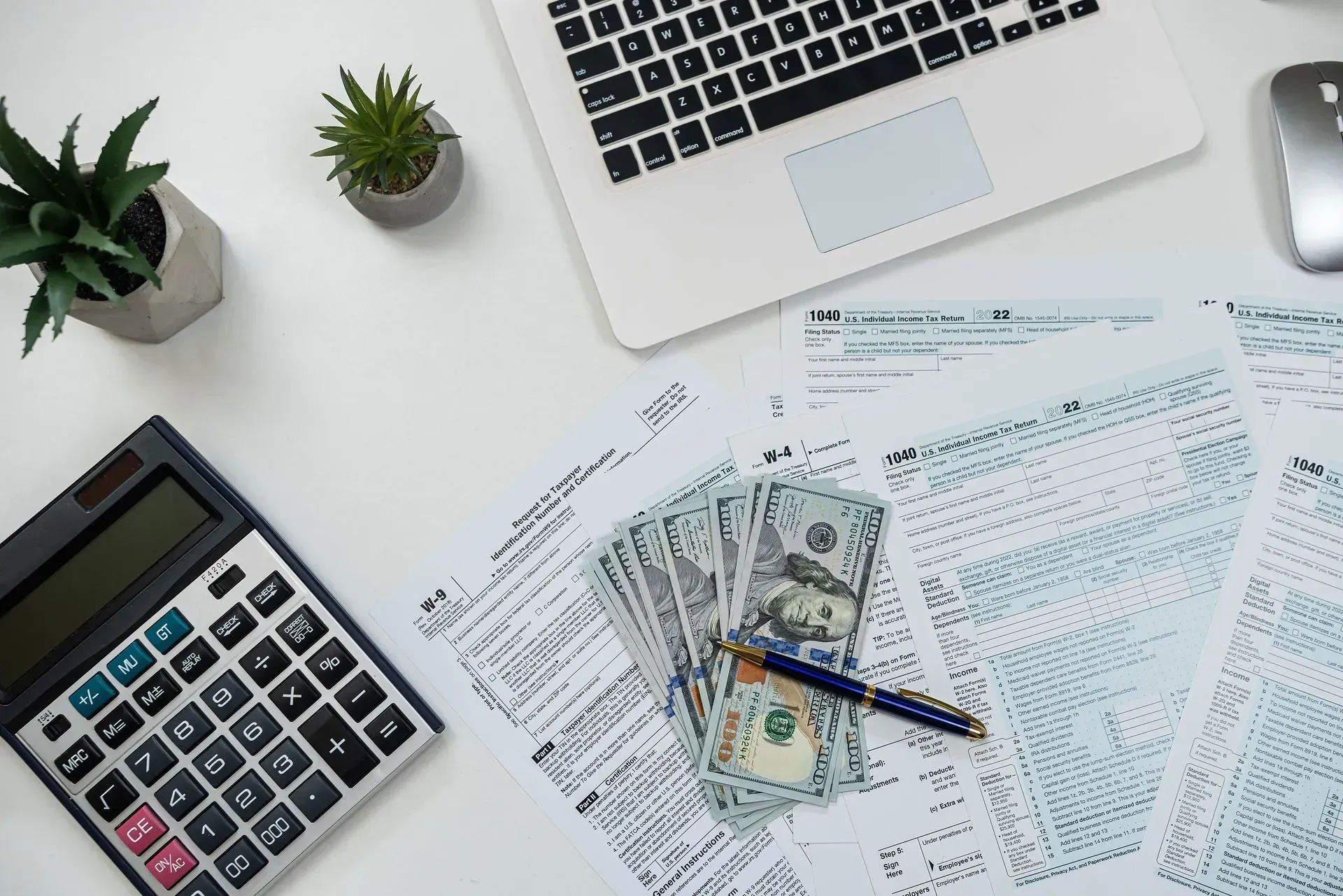 Desk with calculator, laptop, scattered tax forms, cash, and a pen for financial paperwork