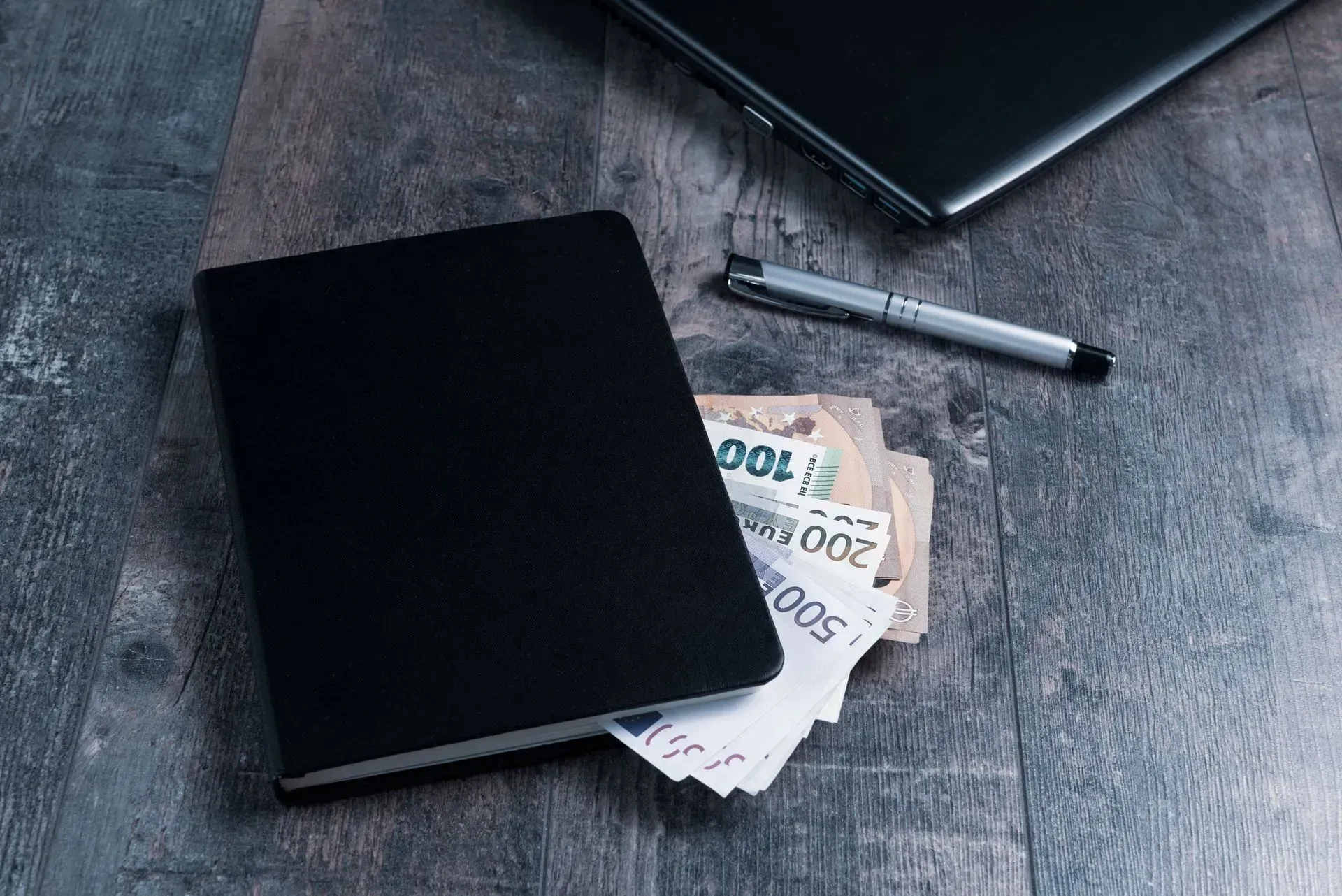 Notebook, pen, and euro banknotes on a gray desk with a laptop corner visible