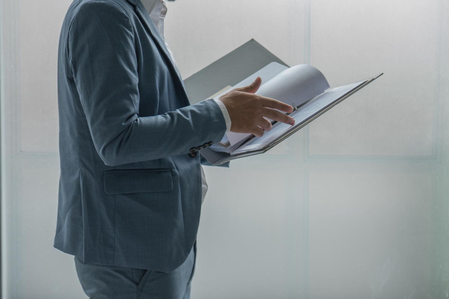 Person in a blue suit reviewing documents in a binder, standing near a bright window.
