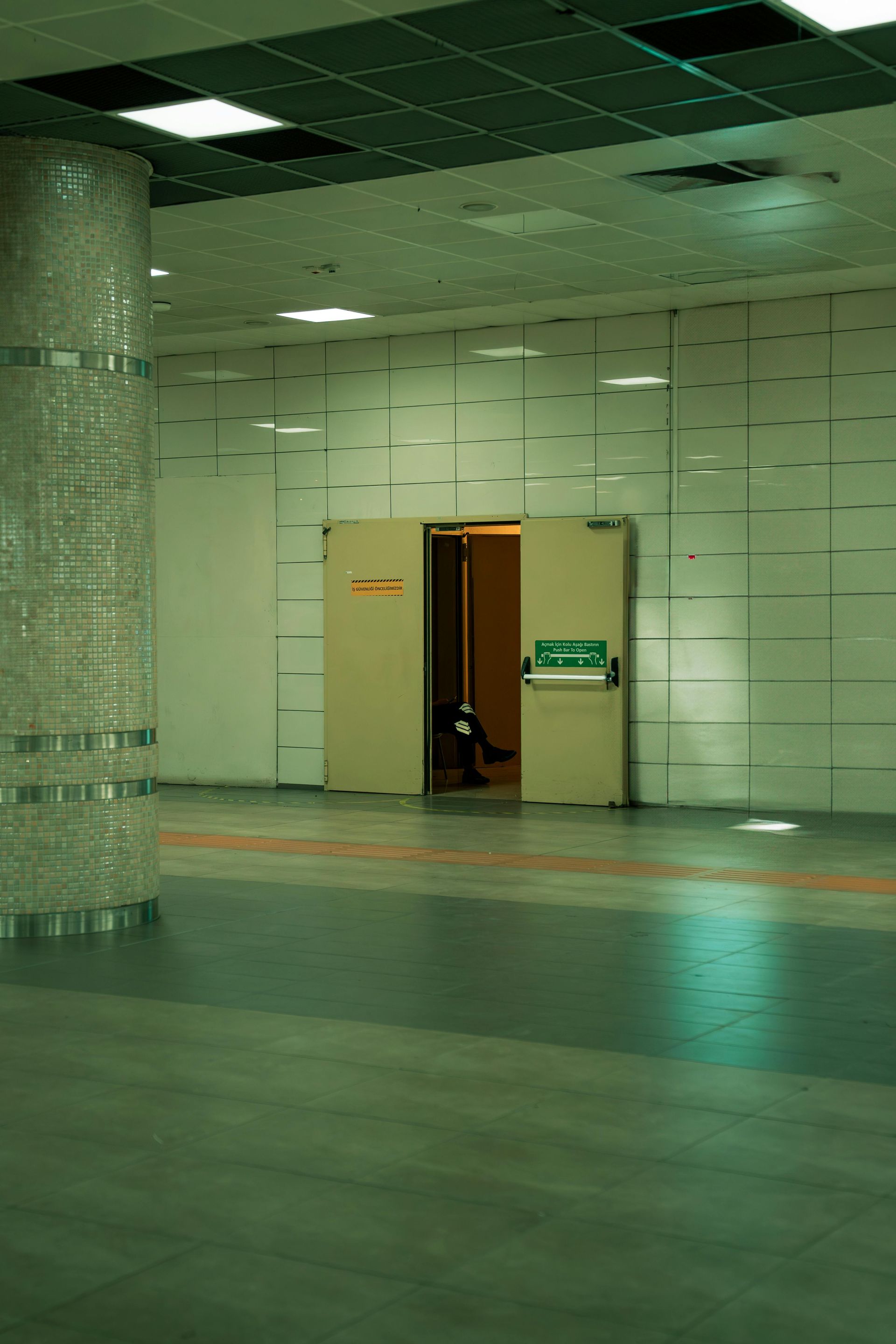 Subway platform with a column on the left and a door with an open interior in the center. Green and gray tones.