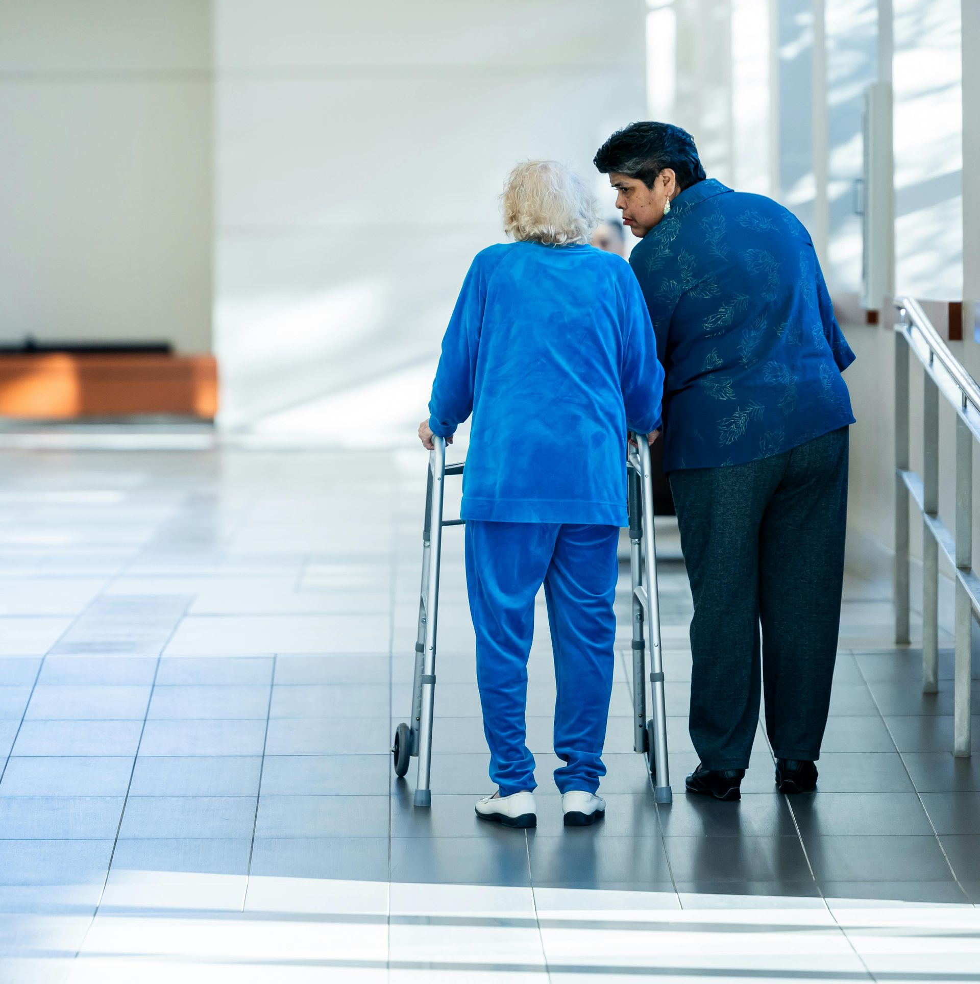Person assisting someone using a walker in a hallway. Both are wearing blue.