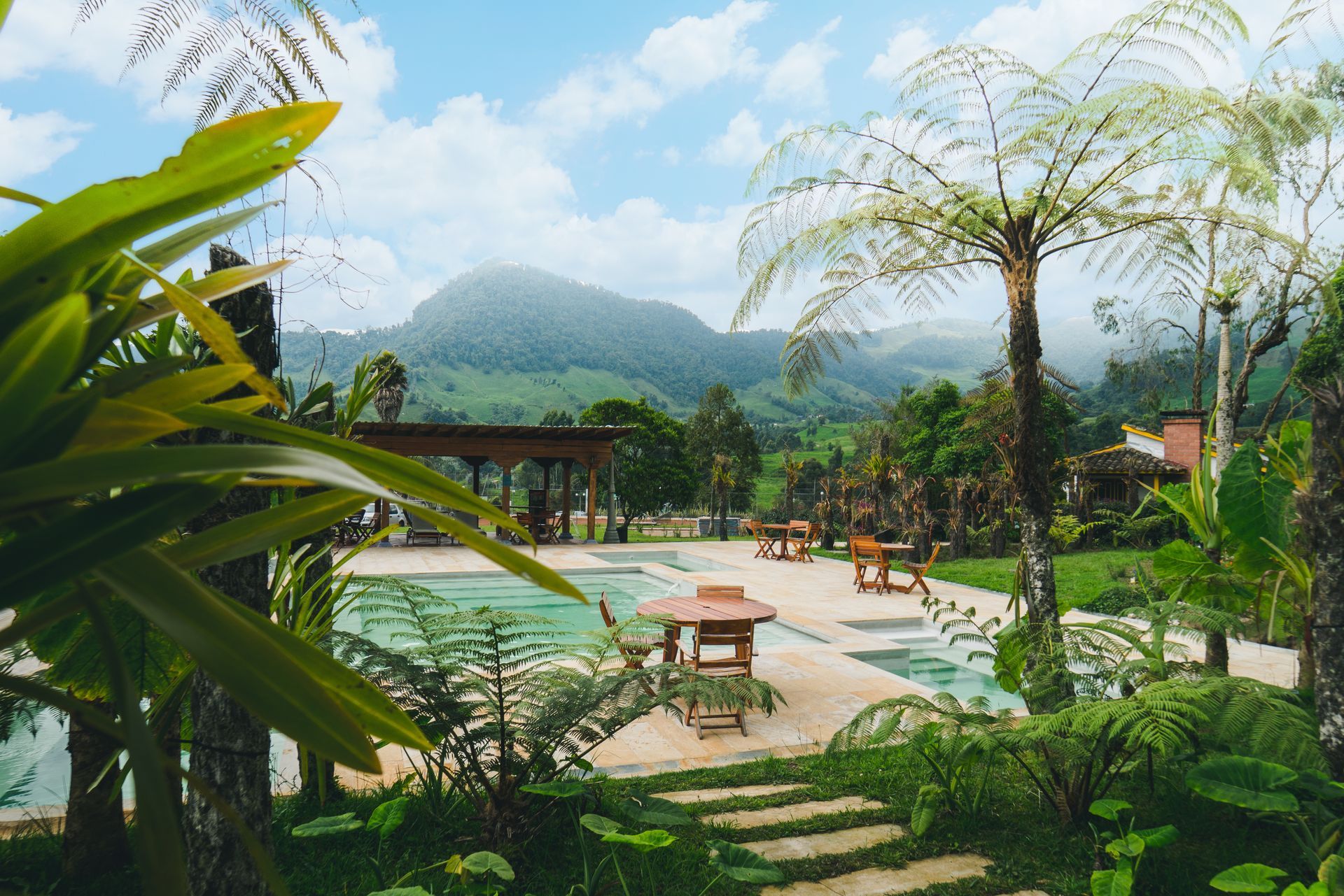 Piscina al aire libre rodeada de jardines, mesas y montaña al fondo en Termales El Otoño.