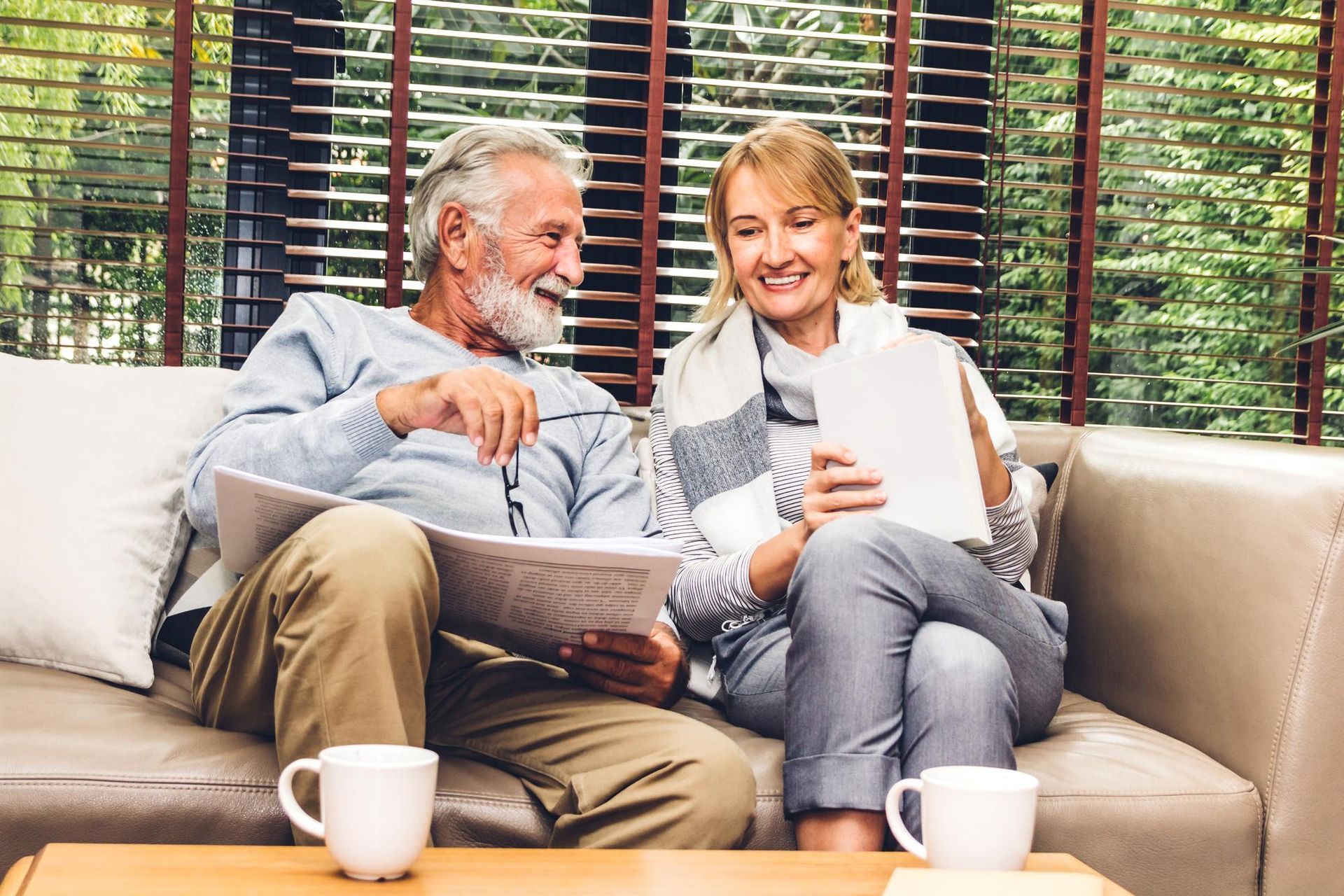 Two people relaxing on a beige couch, smiling and chatting with coffee mugs in a bright living room.