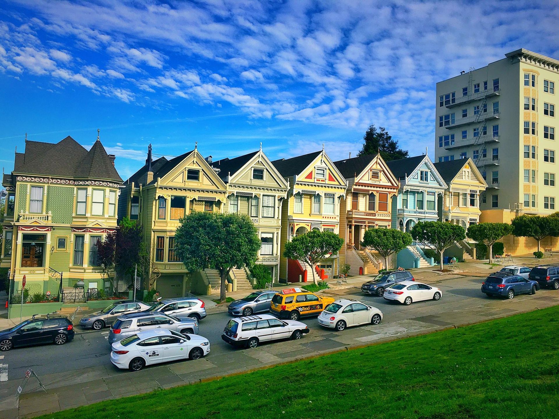 Colorful Victorian houses beside a city street with parked cars, under a bright blue sky