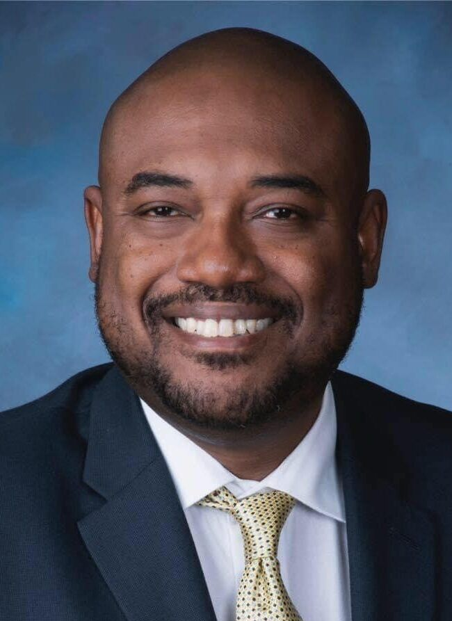 Smiling man in a dark suit and yellow tie against a blue studio background