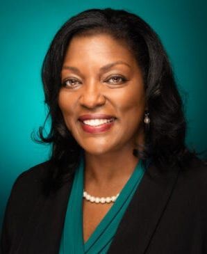 Smiling professional portrait on a teal background, wearing a black blazer and pearl necklace.