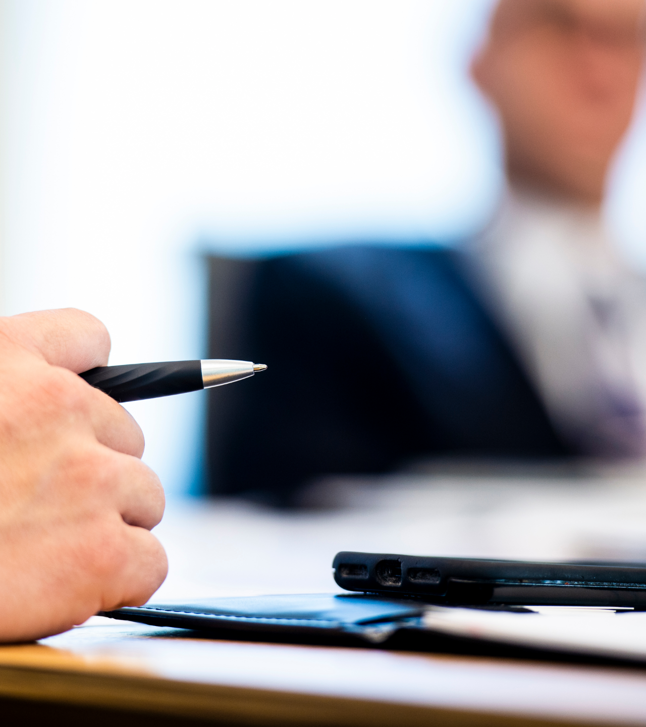 Hand holding a pen over a laptop in a blurred office meeting scene