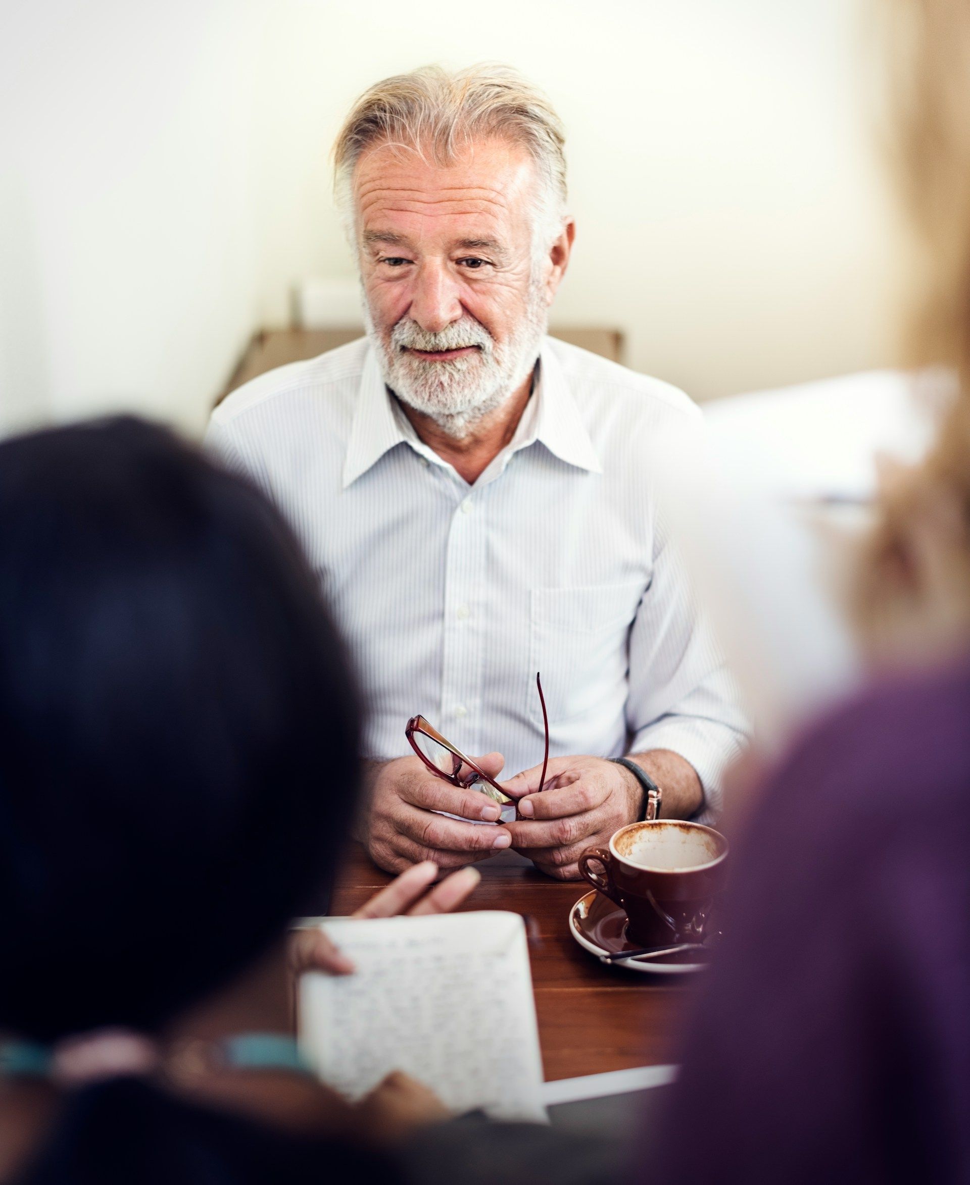 Small group meeting with an older man speaking at a table, holding glasses, in a bright room