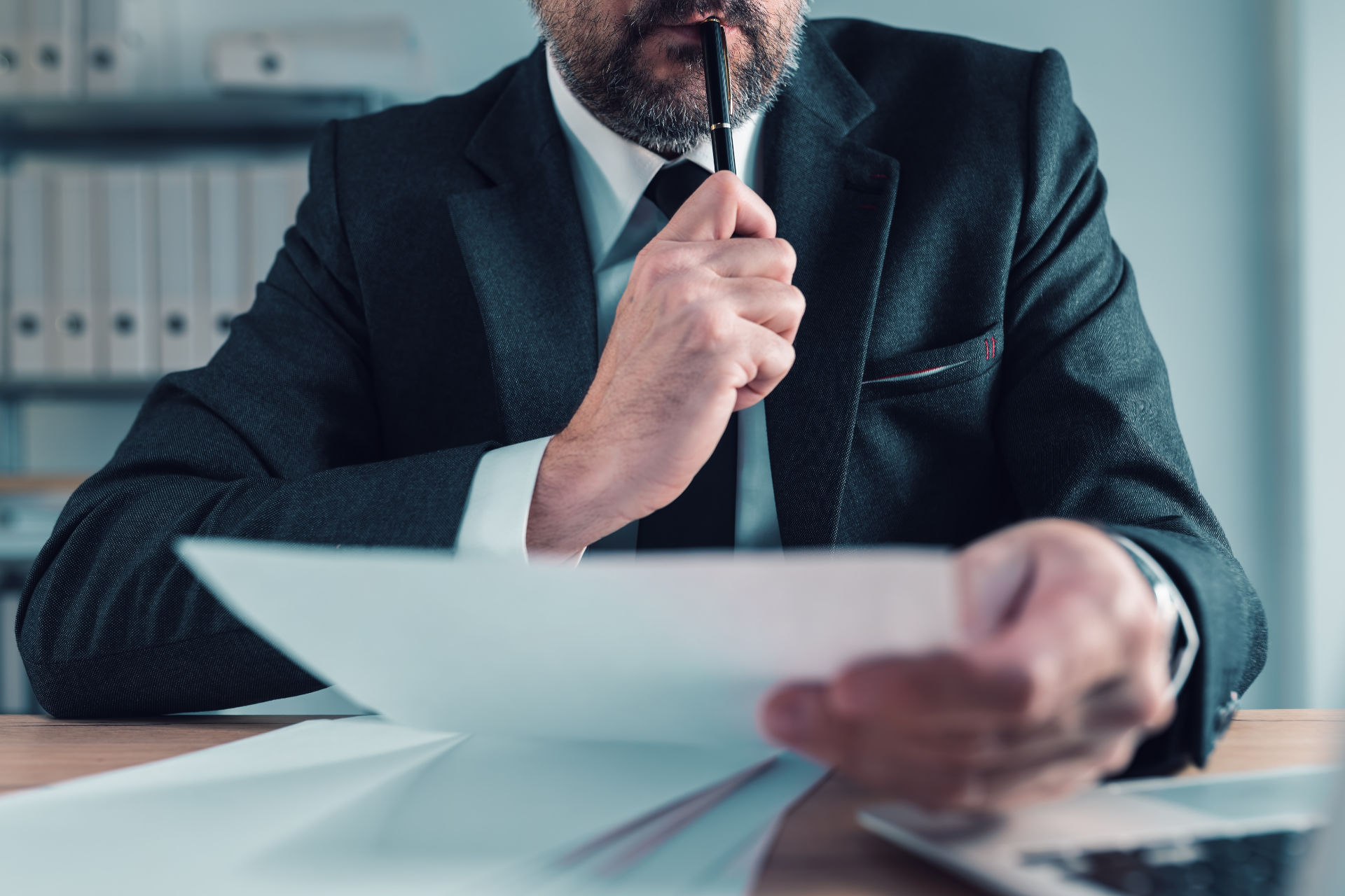 Businessman in suit reviewing documents at a desk, holding papers and a pen.