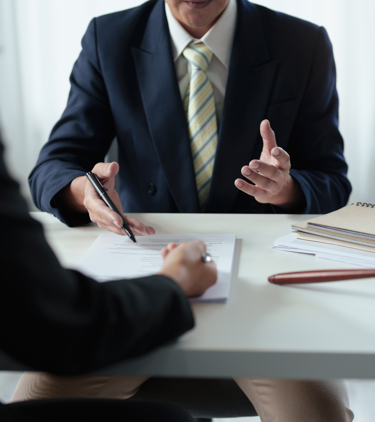 Two people in a business meeting, one gesturing while the other takes notes at a desk.