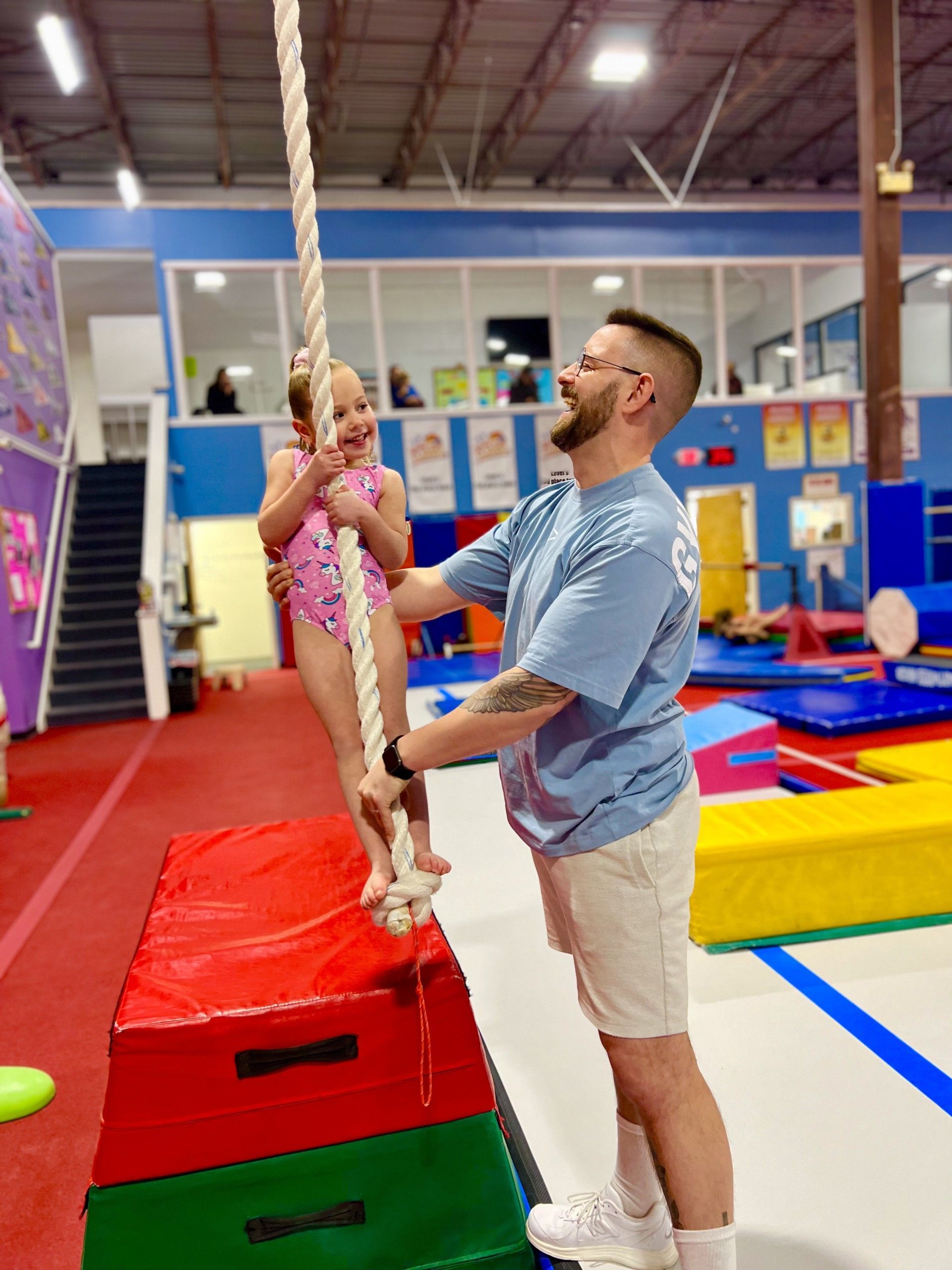 Man helping child climb a rope in a colorful indoor gym, with mats and foam blocks nearby