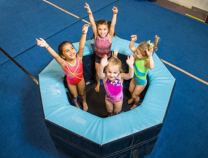 Four children bouncing in a blue trampoline, smiling with arms raised in a gym.