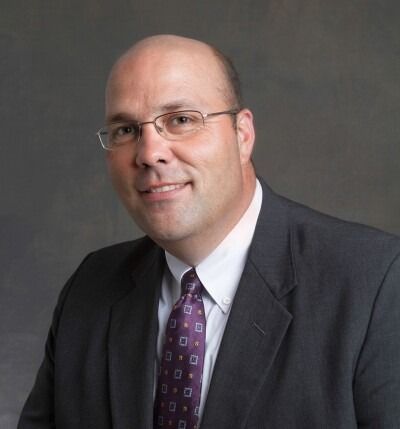 Man in a suit and tie, wearing glasses, smiling slightly, against a dark gray background.