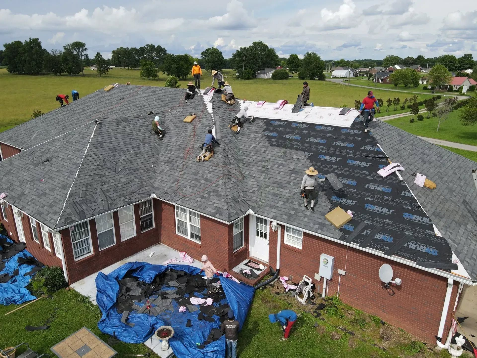 Workers repairing a brick house roof with dark shingles and blue tarps below