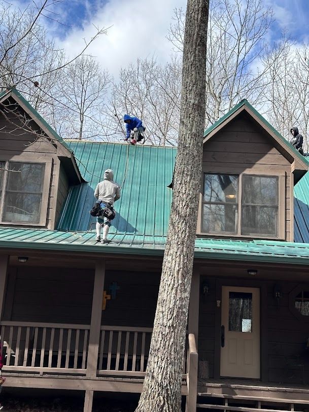 Two workers on a house roof, one in a blue jacket and one in gray, near a tall tree.