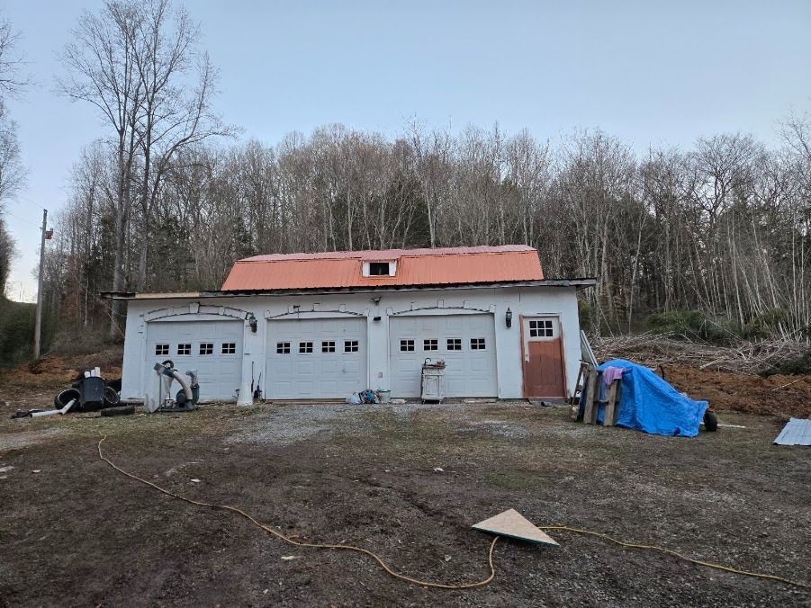 Small garage with red roof in a muddy yard, with a blue tarp and leafless trees behind it.