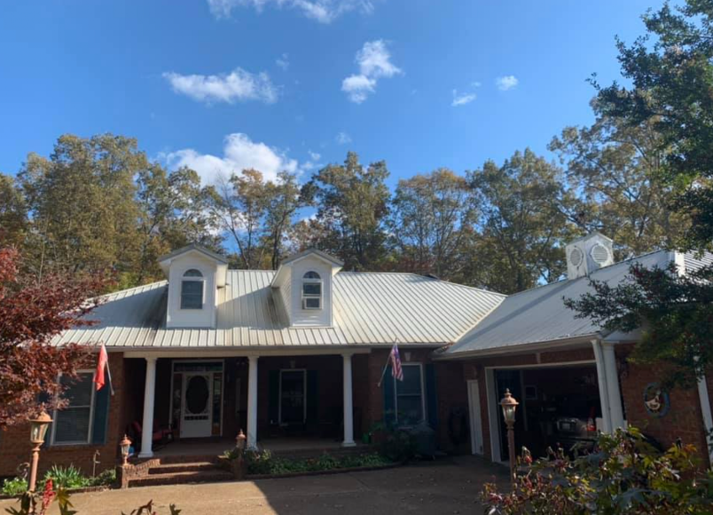 Single-story brick house with white roof and dormer windows, framed by trees under a blue sky.