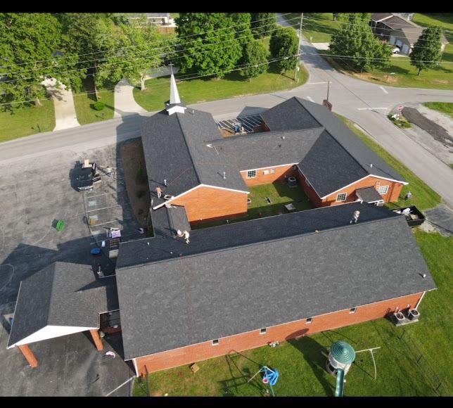 Aerial view of a red-brick building with dark roofs beside a parking lot and trees
