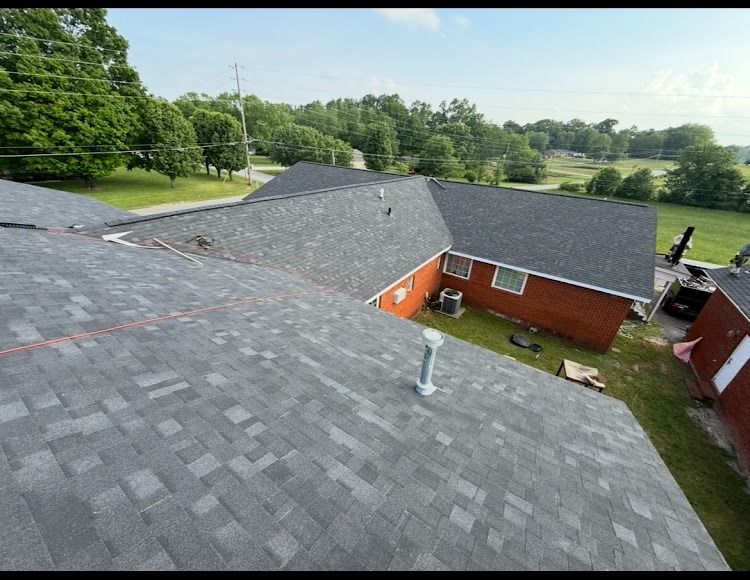 Rooftop view of gray shingle roofs on a red brick building, with trees and open fields in the background.