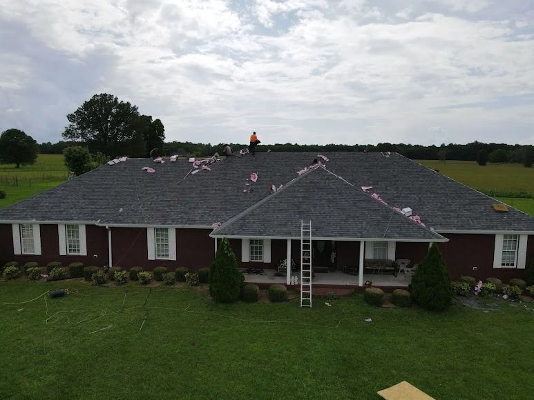 Single-story brick house with damaged roof and workers, one on a ladder, under cloudy skies