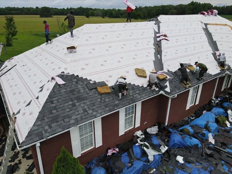 Roofers installing white underlayment and gray shingles on a damaged red-brick house roof