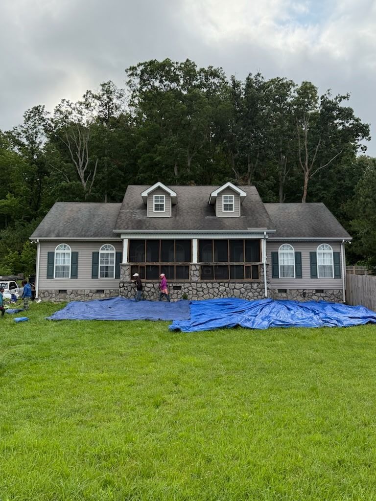 House with a front porch and blue tarp-covered lawn after storm damage, with a person standing on the porch.