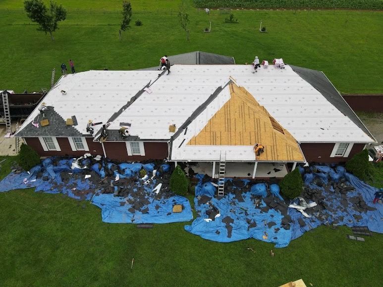 Aerial view of a house with a white roof being repaired, with blue tarps and workers on top.