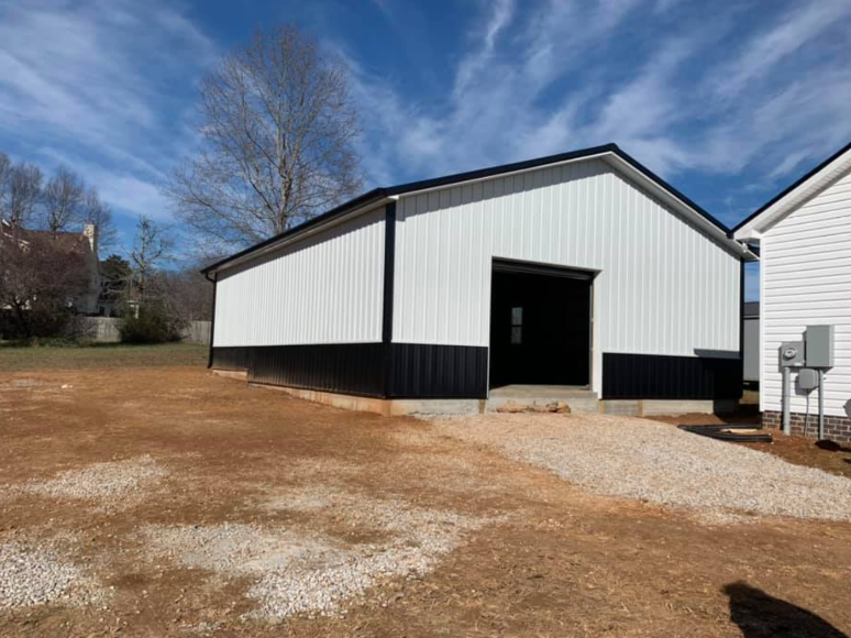 White-and-black metal barn building with an open doorway on a gravel lot under a blue sky.
