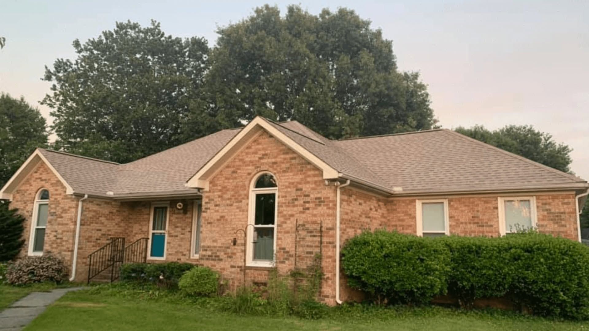 Single-story brick house with a gray roof, arched window, and trimmed hedges under cloudy sky.