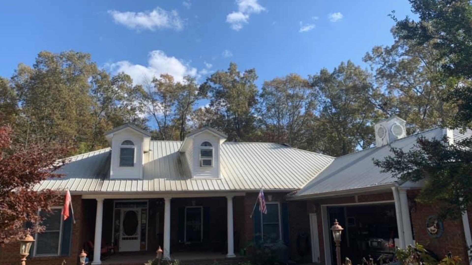 Brick house with white roof and dormers under a blue sky, framed by trees and American flags on the porch