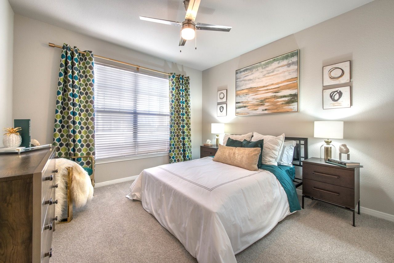 Bedroom in an apartment with a bed, two nightstands, a ceiling fan, and a large window with curtains.