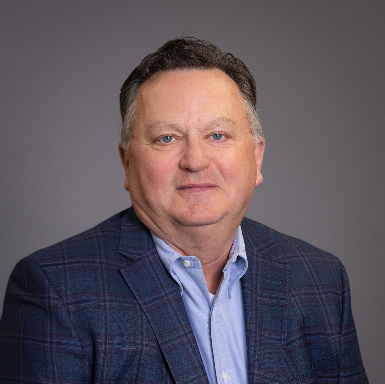 A man in a suit and blue shirt is sitting in front of a gray background.
