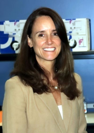 A woman in a tan jacket is smiling in front of a blue wall with the letter f on it