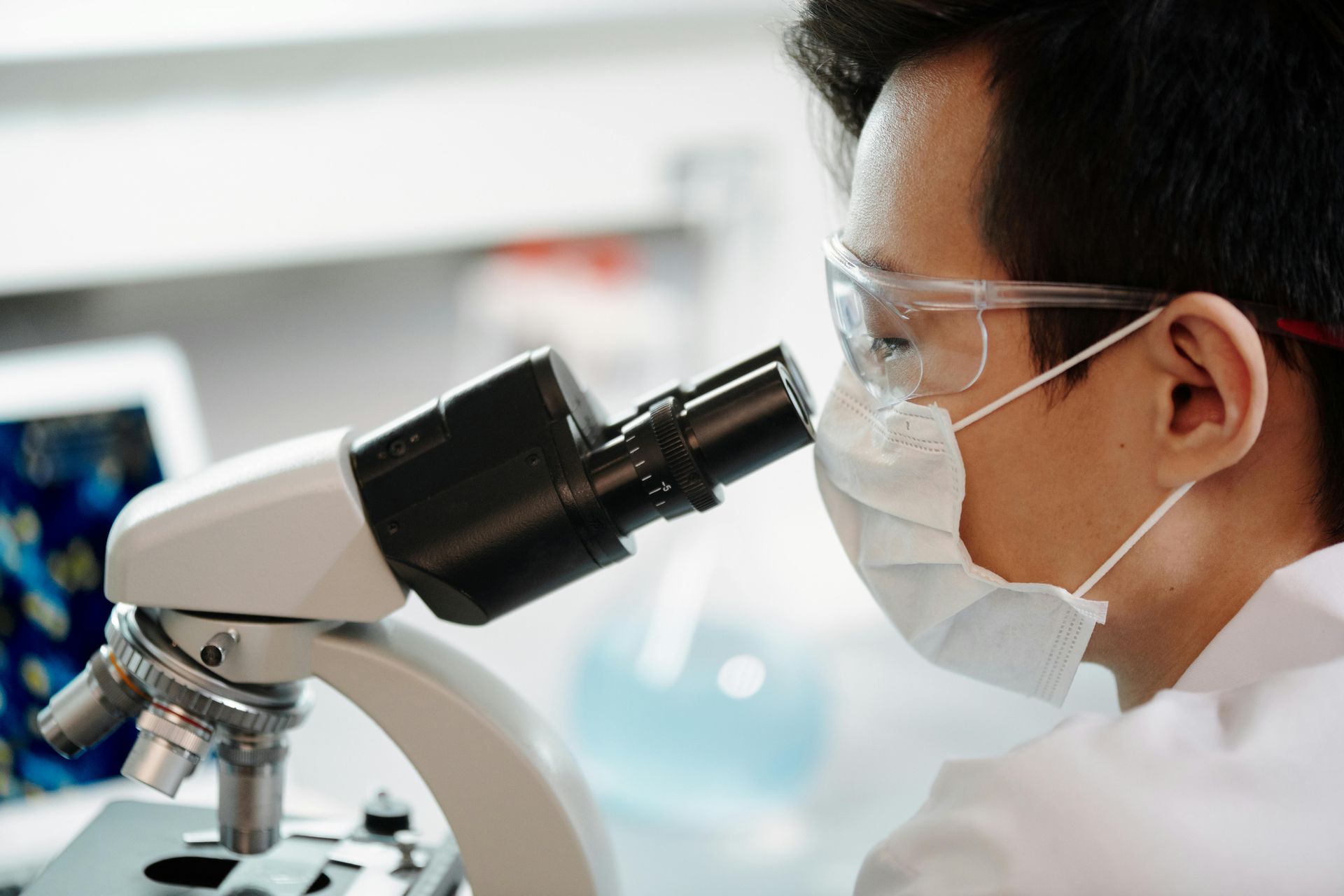 A man wearing a mask and goggles is looking through a microscope.