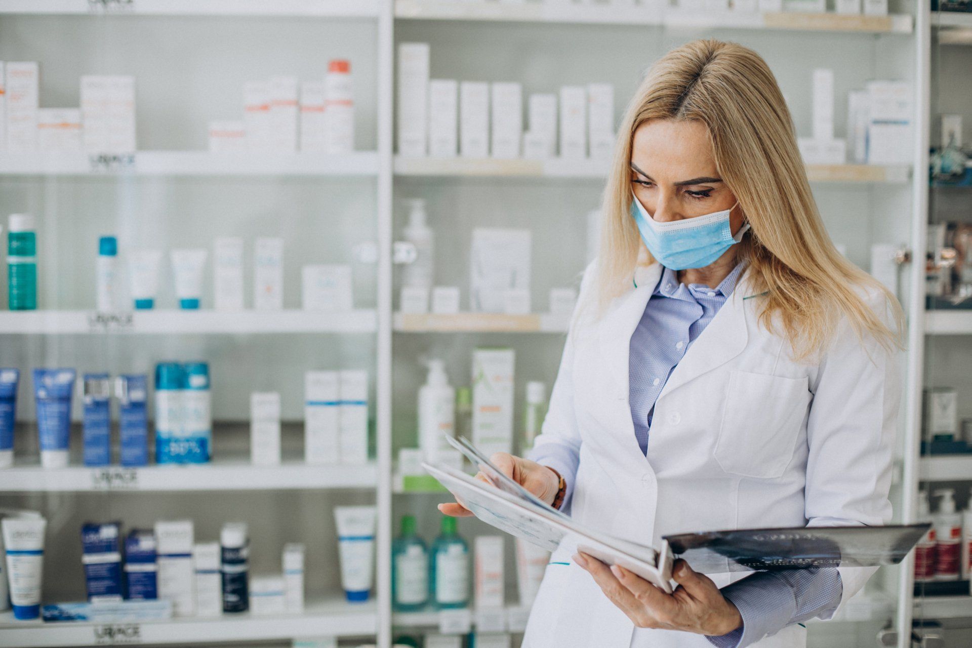 A female pharmacist wearing a mask is looking at a clipboard in a pharmacy.