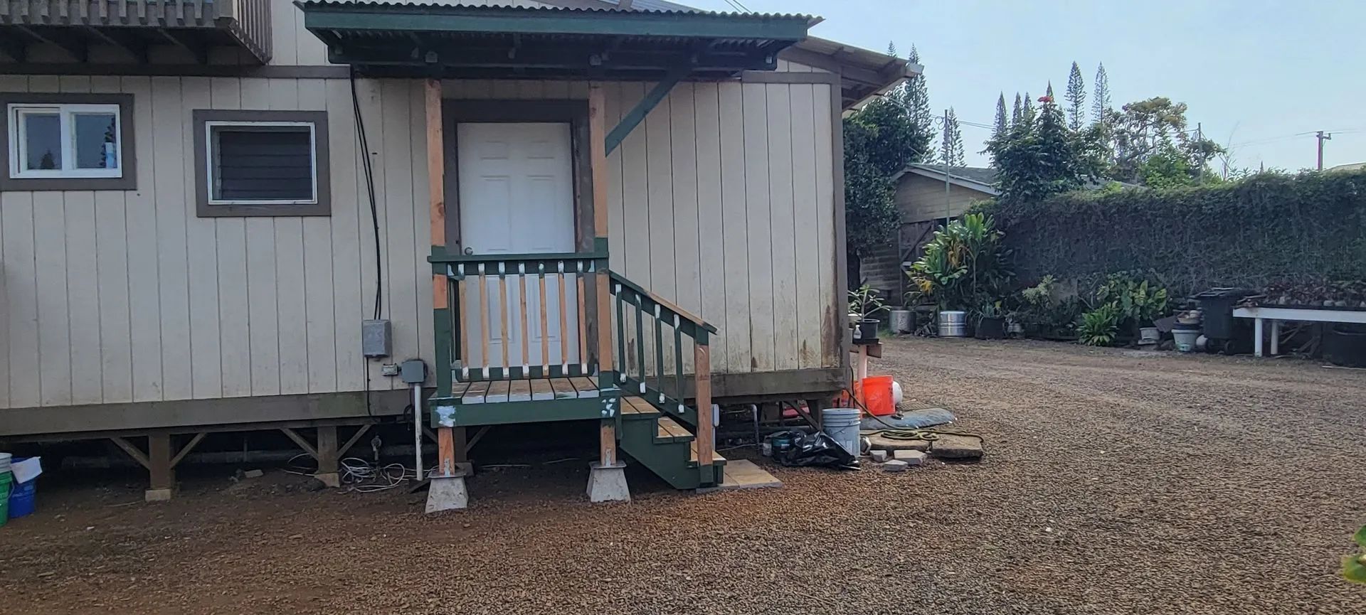 Small beige shed with a front porch and steps on a gravel lot, with trees and a bench nearby.