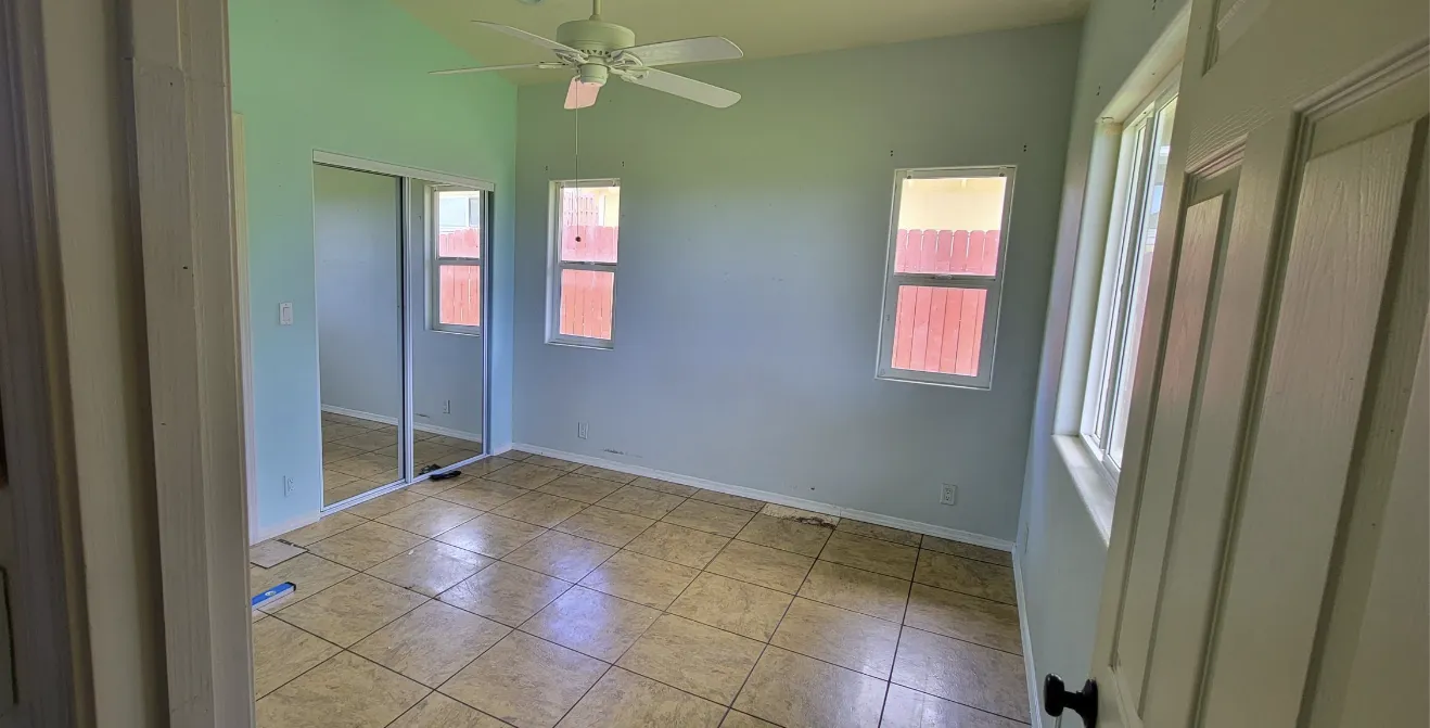 Empty bedroom with pale green walls, tiled floor, ceiling fan, mirrored closet doors, and two windows.