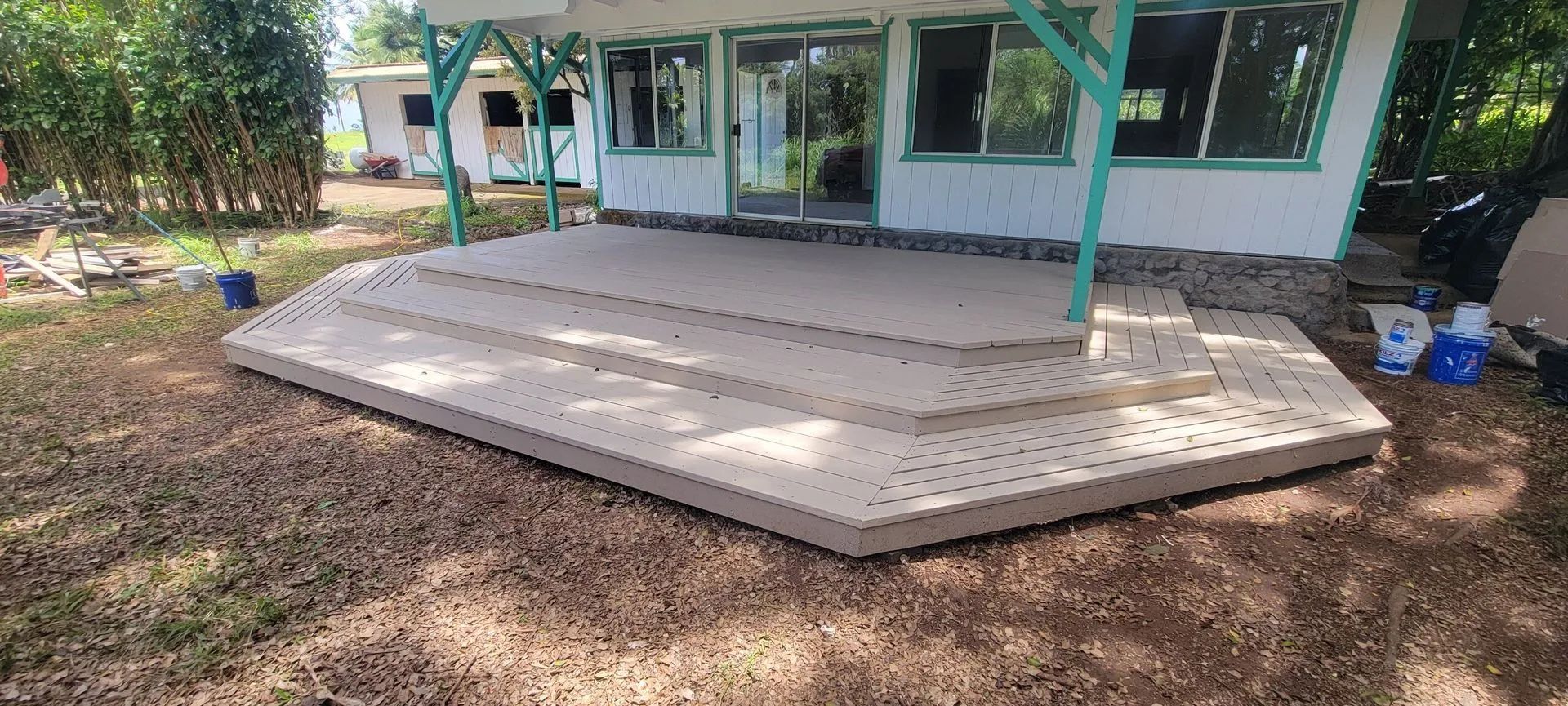 Wide wooden deck extension beside a house, with green trim and dirt yard around it.