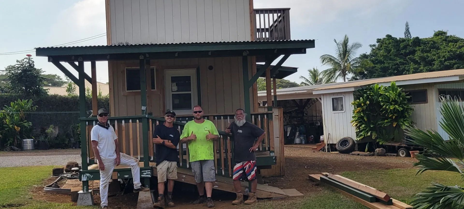 Five people stand in front of a small wooden house under construction with tools and materials nearby