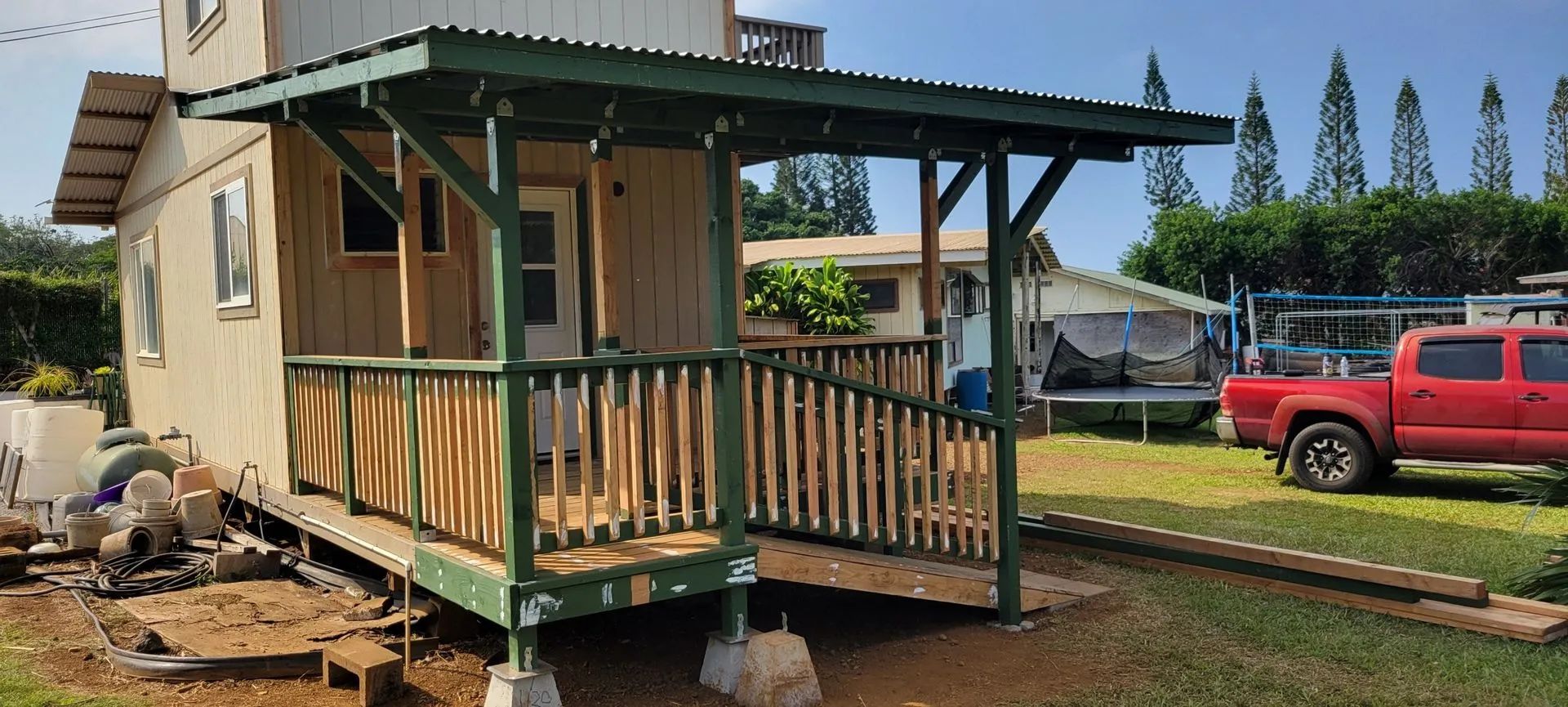 Small beige cabin with wooden porch on jacks in a yard, with a red pickup truck nearby