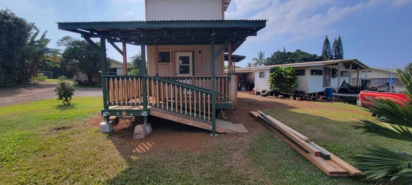 Small wooden porch on a tan building with a ramp, in a grassy yard under bright sun.