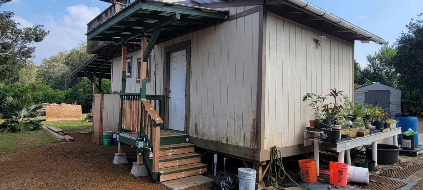 Small white house with a porch, steps, potted plants, and a wet yard after rain