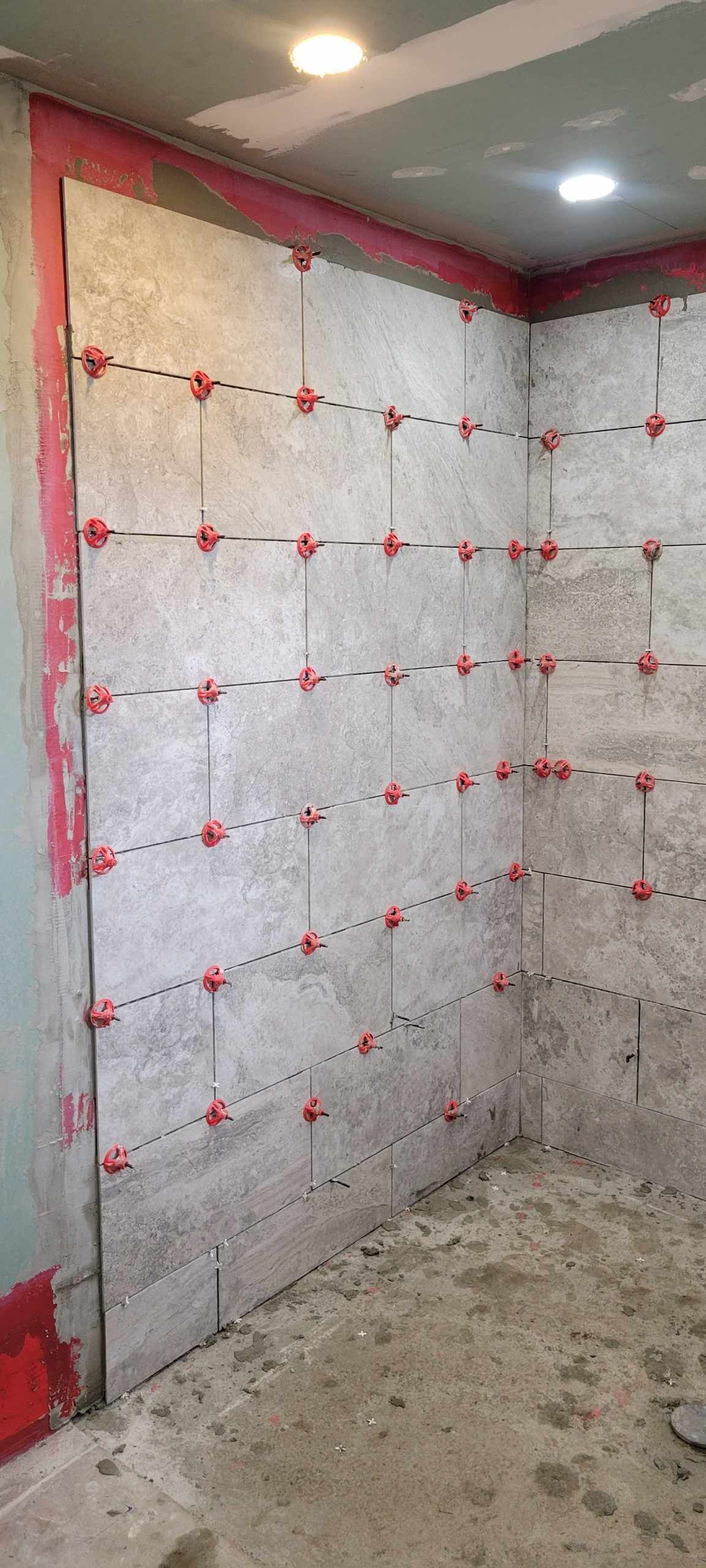 Tiled shower corner with red leveling clips, red waterproofing edges, and unfinished concrete floor