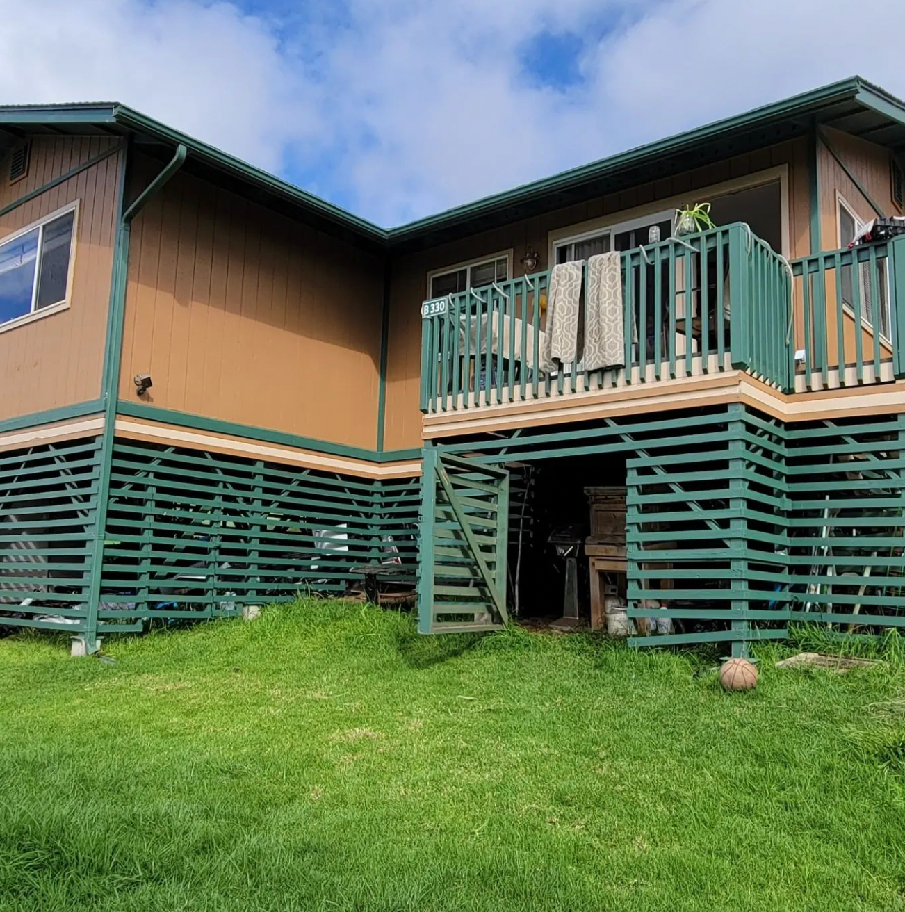 Stilt house with teal railings and stairs on a grassy hillside under a partly cloudy sky